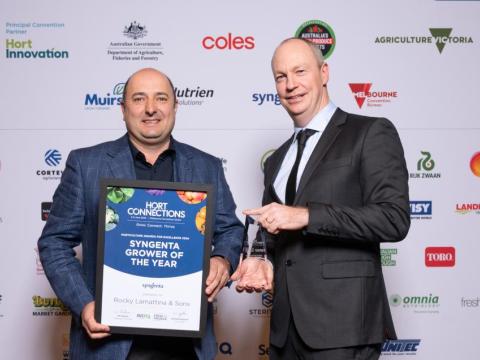 Two men shaking hands at an awards backdrop, one holding a framed certificate