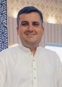 Smiling man in a white traditional shirt indoors, patterned wall behind him