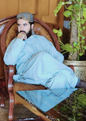 Man in light blue robe reclining in wooden chair beside leafy plants indoors