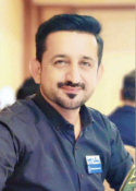 Smiling man in a dark shirt with a name badge, seated indoors with blurred background