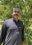 Man with mustache in a dark shirt standing outdoors among green trees