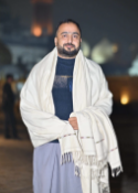 Man in a shawl holding a scarf at night, with blurred city lights behind him.