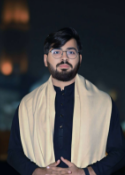 Man with glasses and beard, wearing a cream shawl over black clothing, blurred background.