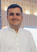 Smiling man in a white traditional shirt indoors, patterned wall behind him