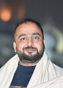 Man in a shawl holding a scarf at night, with blurred city lights behind him.