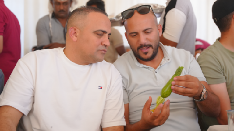 Two men in white shirts examine green seedlings in a crowded room.