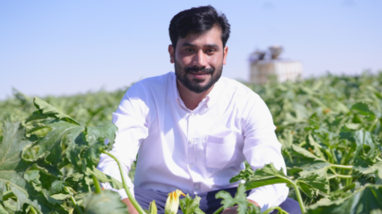 Smiling man in a white shirt crouching in a green crop field under blue sky