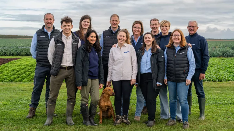 Group of 12 people posing outdoors in a green field with a brown dog