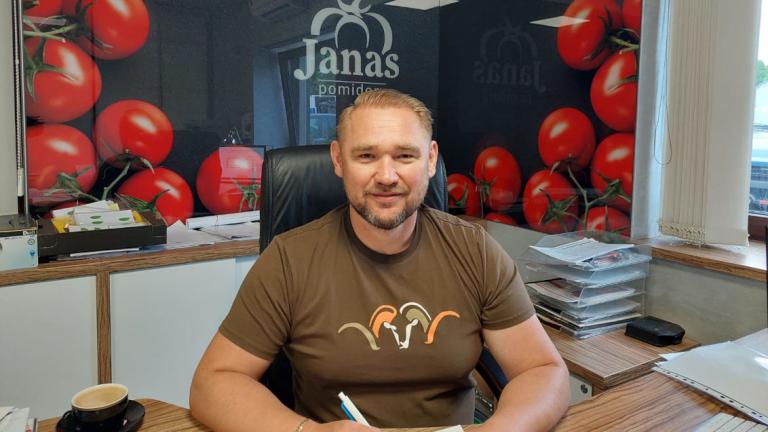 Man in a brown T-shirt sitting at a desk in an office with red balloons behind him.