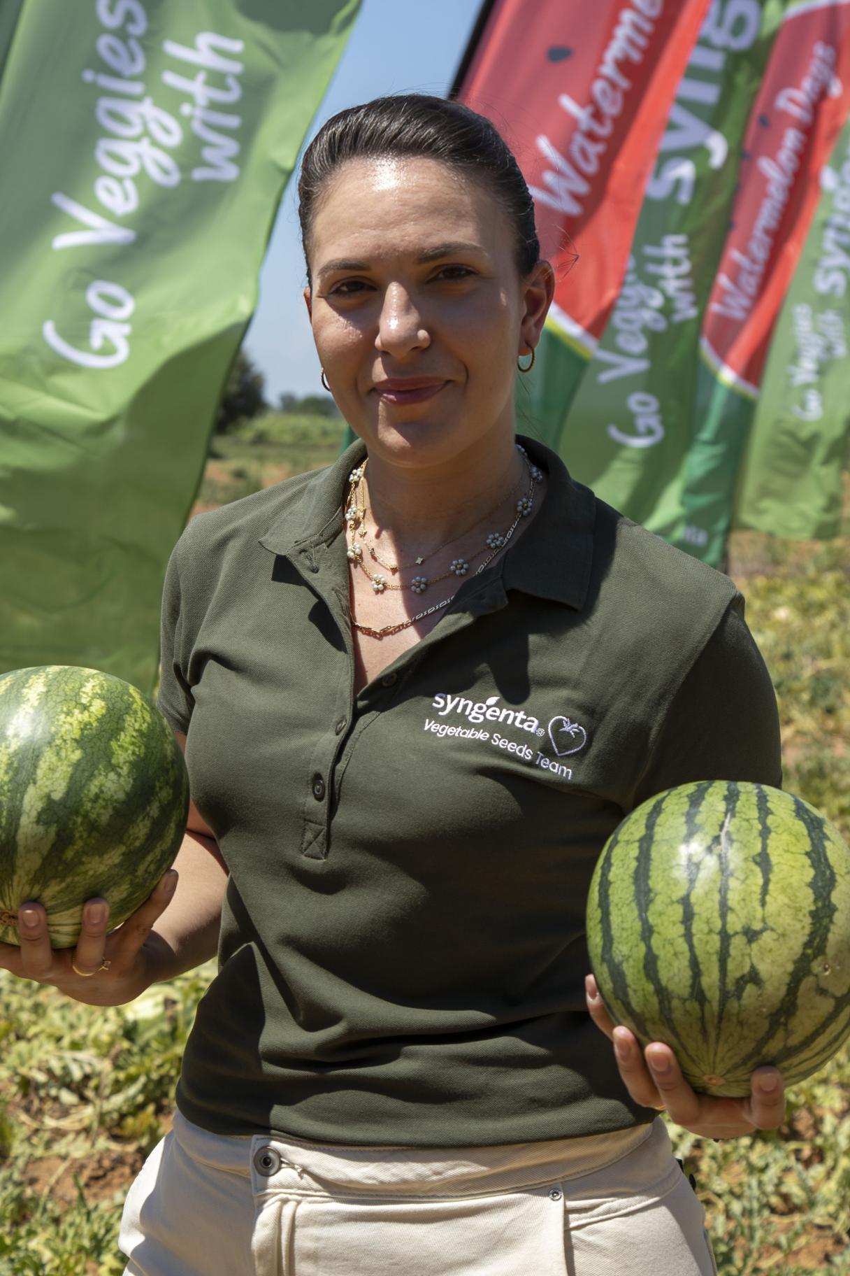 Woman in green polo holding two watermelons in a field with colorful flags