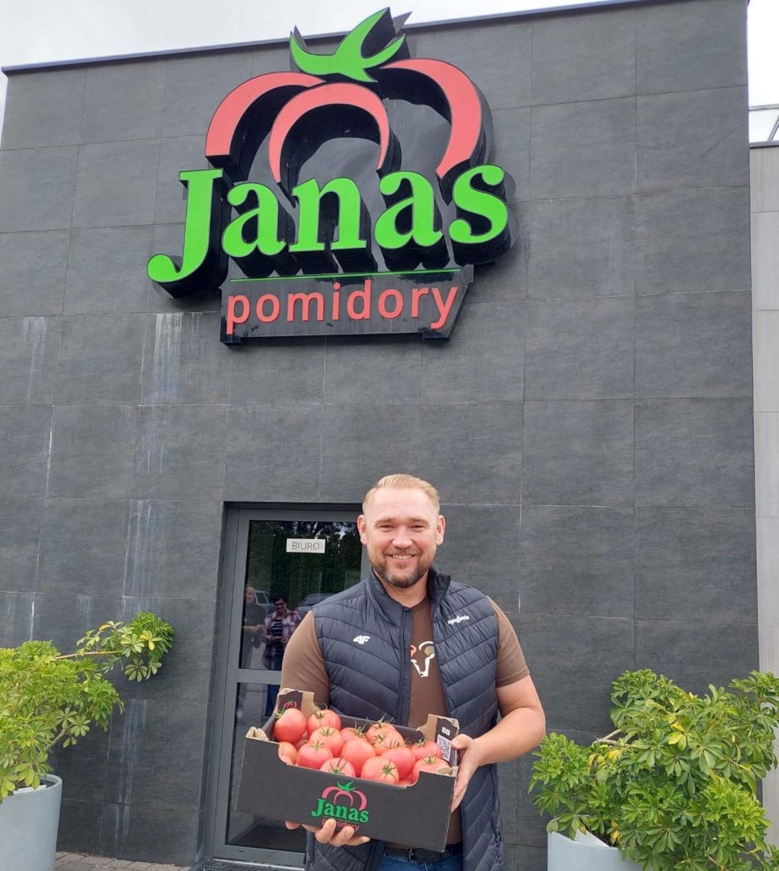Smiling man holding a crate of tomatoes in front of Janas Pomidory storefront sign