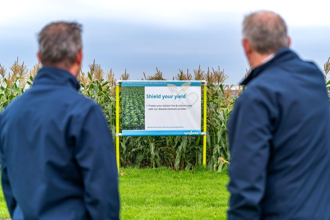 Two men in blue jackets viewing a sign in a green field with crops